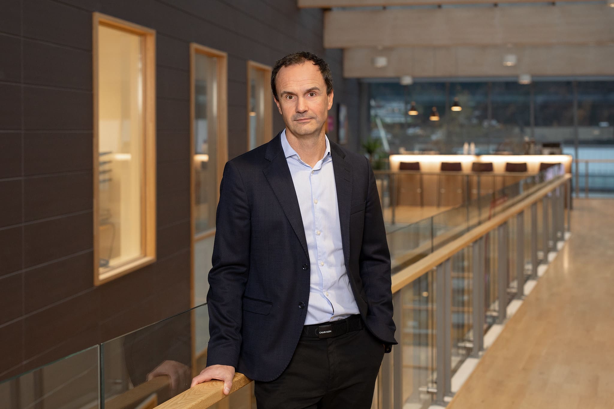 Bilde av Tore Bell, som er avdelingsdirekt&oslash;r i Lotteritilsynet. Han har p&aring; seg svart dress og en l&aring;s bl&aring; skjorte, og st&aring;r ved et gelender inne i lokalene til Lotteri- og stiftelsestilsynet.